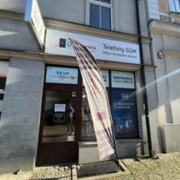 Fasada budynku z szyldem 'Telefony GSM skup-sprzedaż-serwis' i flagą reklamową z napisem 'Telefony Tablety Komputery' oraz informacją o naprawie telefon&oacute;w i skupie telefon&oacute;w w oknach.