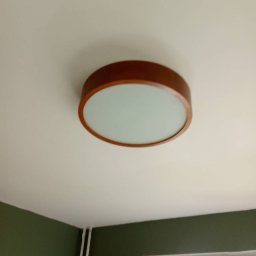 Kamil krzych - Zamontowana okrągła lampa sufitowa z drewnianą obudową i mlecznym kloszem, widoczna na tle białego sufitu i fragmentu zielonej ściany.