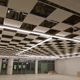 Kamil krzych - Sufit podwieszany kasetonowy z widocznymi elementami instalacji i oświetleniem liniowym LED w nowym lokalu handlowym. Widoczna drabina i materiały budowlane.