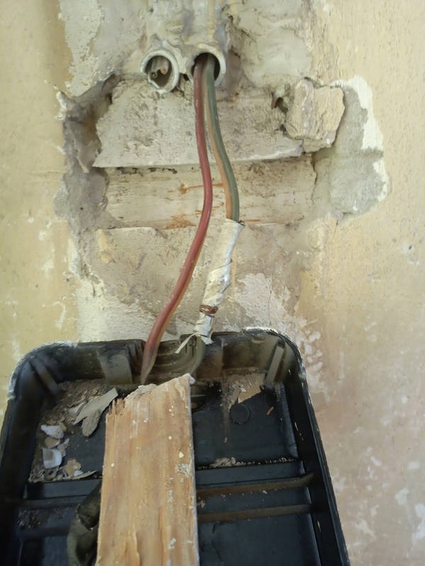 Odsłonięte przewody elektryczne wystające ze ściany z uszkodzonym tynkiem, widoczna puszka instalacyjna z fragmentem deski. Prace remontowe w trakcie realizacji.