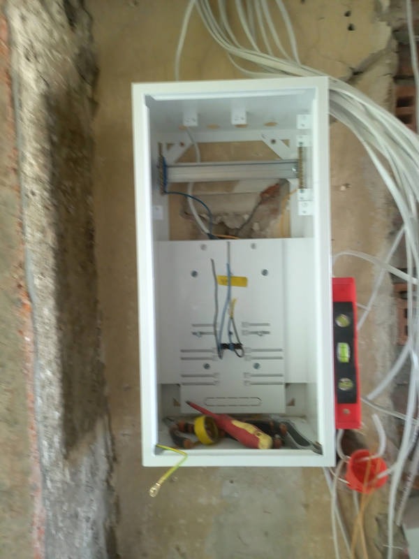 Biała skrzynka rozdzielcza w trakcie montażu na surowej ścianie, widoczne przewody, poziomica i narzędzia. Instalacja elektryczna w trakcie realizacji.