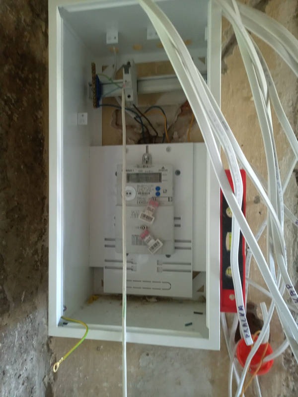 Biała skrzynka z licznikiem elektrycznym na ścianie, widoczne kable i poziomica. Instalacja w trakcie montażu, surowa ściana w tle.
