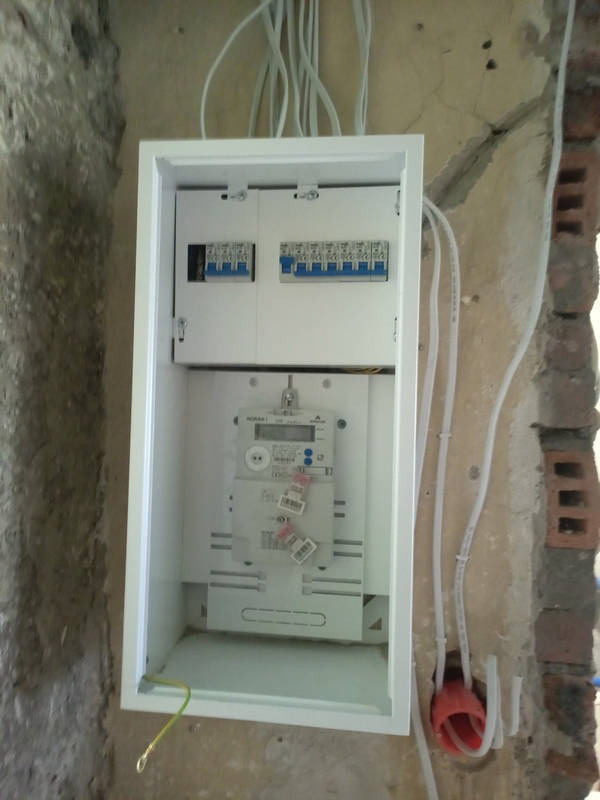 Biała skrzynka z zamontowanym licznikiem i bezpiecznikami na surowej ścianie z widocznymi przewodami. Instalacja w trakcie prac wykończeniowych.