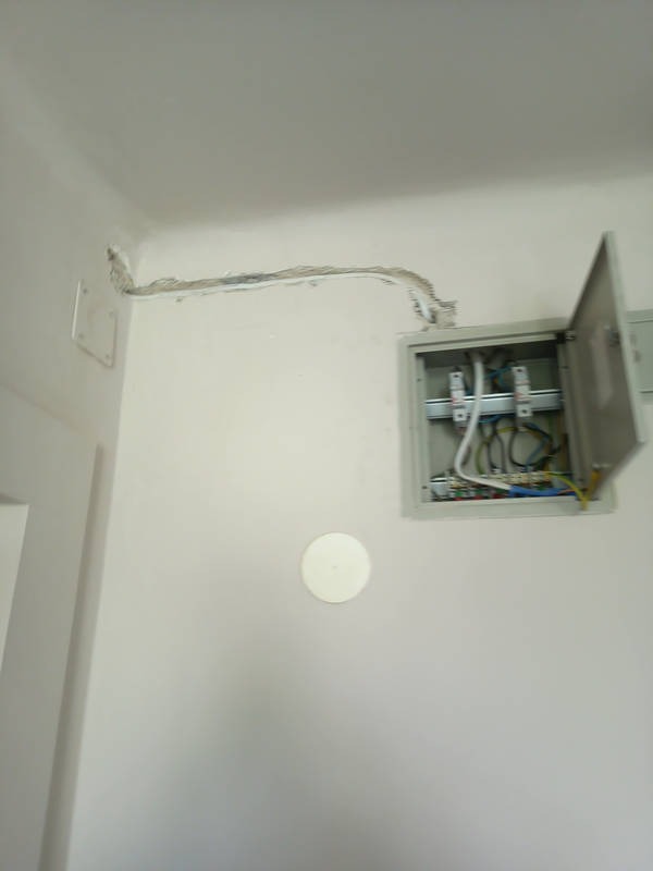 Otwarta skrzynka elektryczna na białej ścianie z wystającymi kablami, widoczne przewody w ścianie. Białe tło, surowy stan wykończenia.