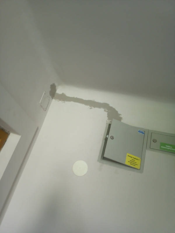 Otwarta skrzynka elektryczna na ścianie, widoczne ślady po naprawie kabli. Biała ściana i sufit, widoczne kable w szarej masie naprawczej.