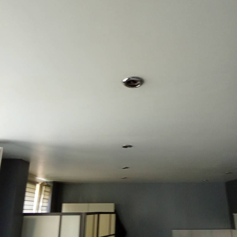 Równomiernie rozmieszczone, okrągłe, metalowe oprawy oświetleniowe w białym suficie, widoczne z dołu, w tle szara ściana i okno z żaluzjami.