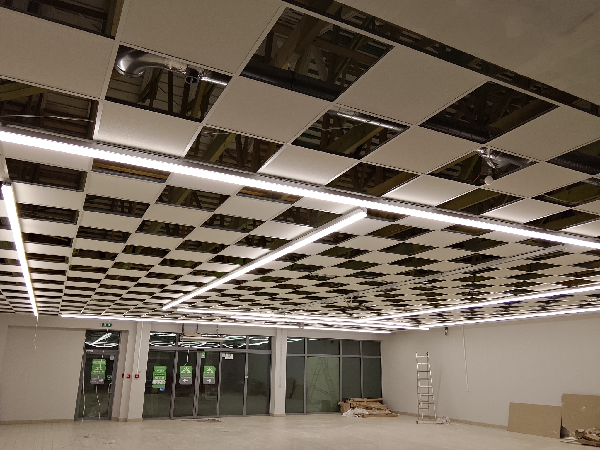 Sufit podwieszany kasetonowy z widocznymi elementami instalacji i oświetleniem liniowym LED w nowym lokalu handlowym. Widoczna drabina i materiały budowlane.