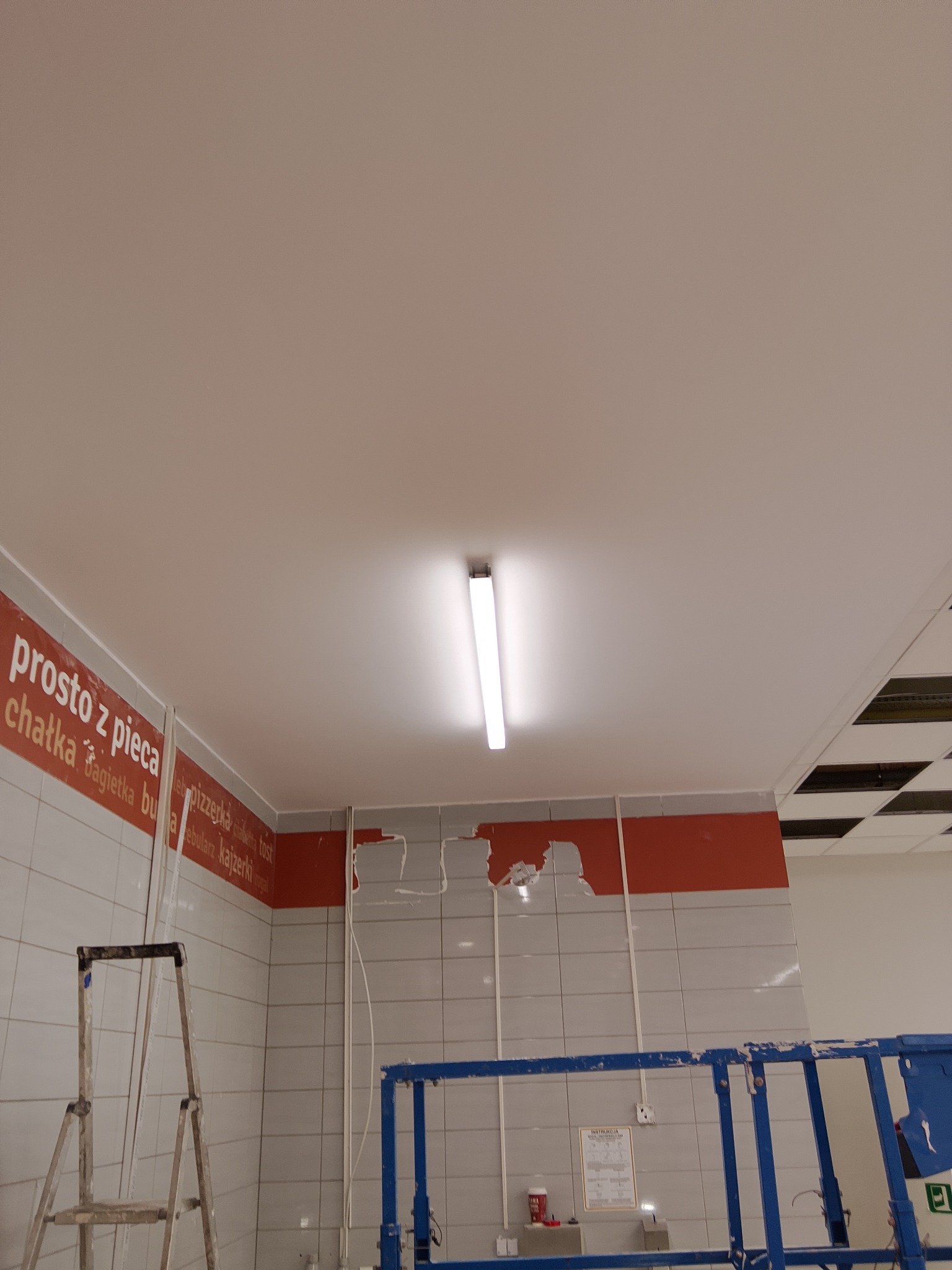 Remont lokalu gastronomicznego: uszkodzona ściana z płytek, widoczna instalacja elektryczna, drabina i podnośnik. Przygotowanie do montażu oświetlenia.
