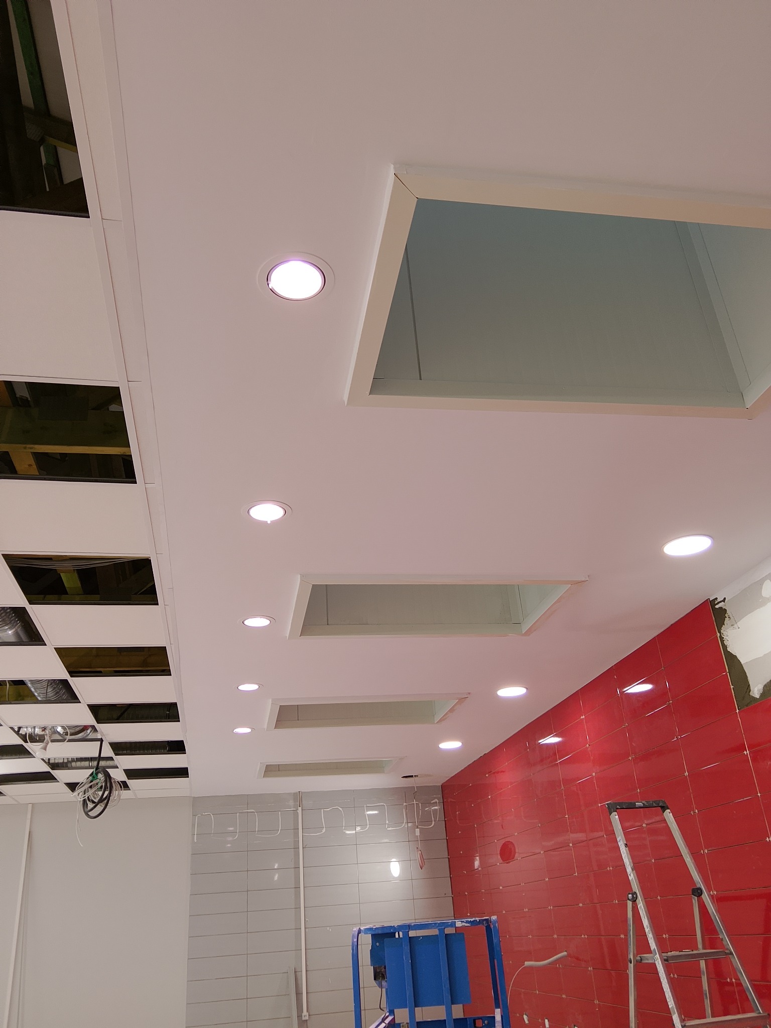 Biały sufit podwieszany z zamontowanymi okrągłymi lampami LED, widoczne otwory i częściowo wykończona ściana z czerwonymi płytkami. W tle drabina i podnośnik.