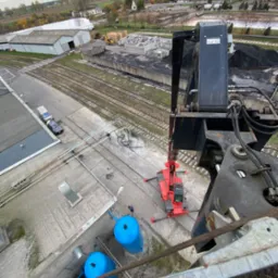 Widok z góry na czerwoną miniładowarkę na betonowym placu, obok niebieskie zbiorniki, w tle budynki przemysłowe, torowisko i wieś.