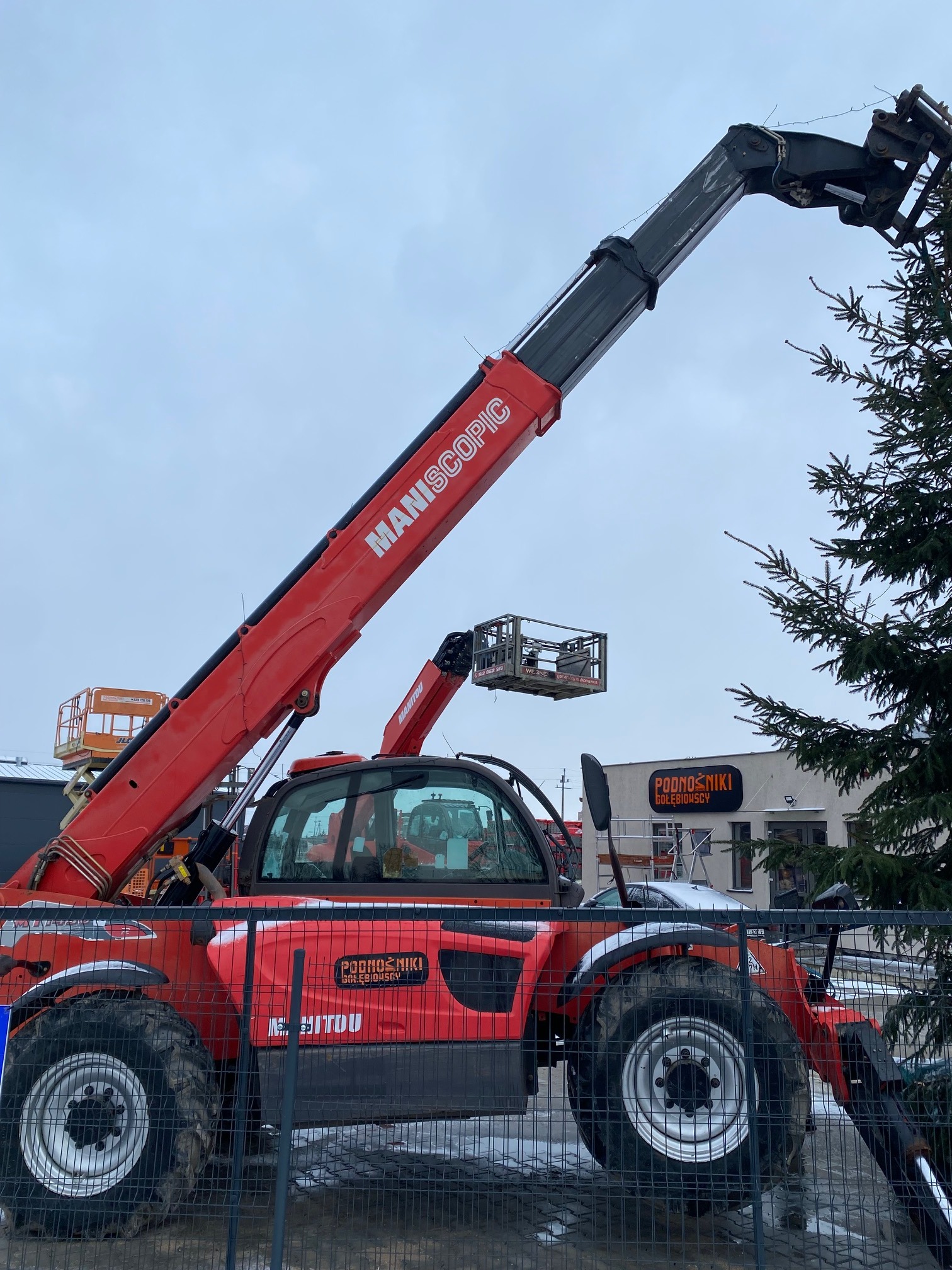 Czerwona ładowarka teleskopowa Manitou z podnośnikiem koszowym na tle pochmurnego nieba i budynku z napisem 'Podnośniki Gołębiowscy', widziana przez metalowe ogrodzenie.