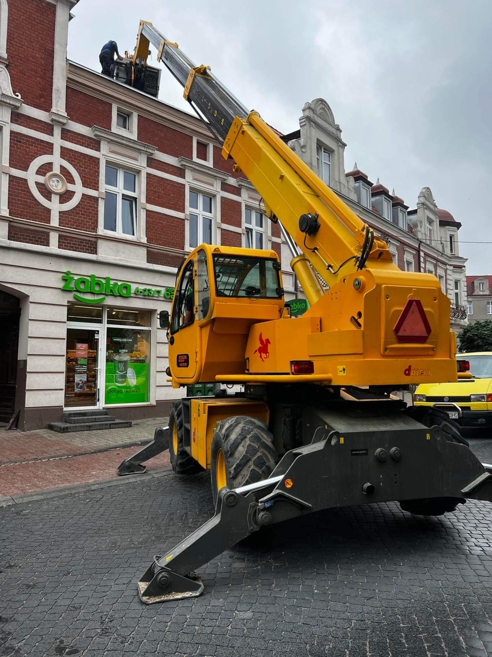 Żółty podnośnik koszowy z wysięgnikiem unosi pracownika na dach budynku z szyldem Żabka, widoczne podpory i kostka brukowa.