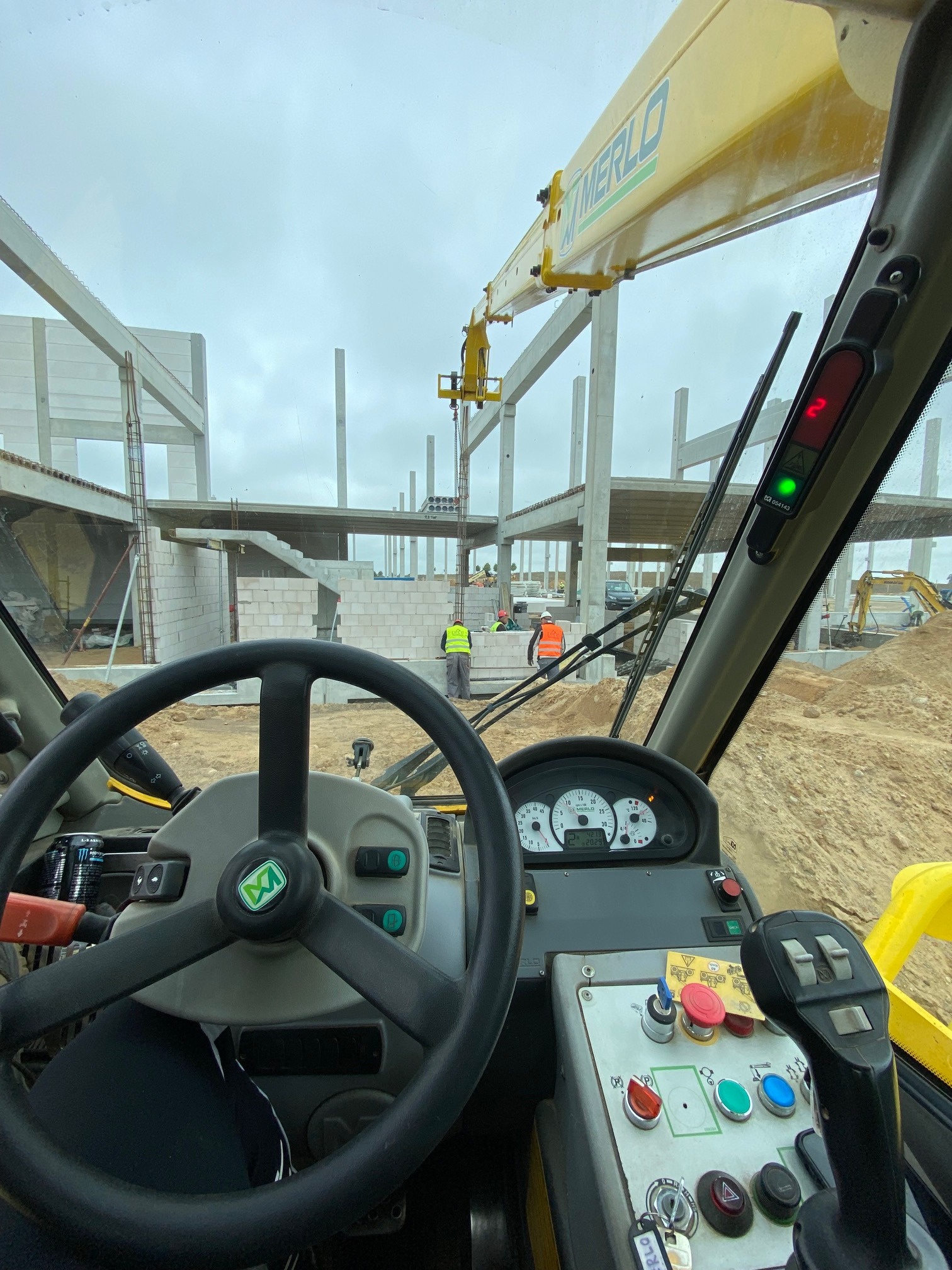 Widok z kabiny żółtego podnośnika teleskopowego Merlo na plac budowy z pracownikami w kamizelkach odblaskowych i niedokończoną konstrukcją budynku w tle.