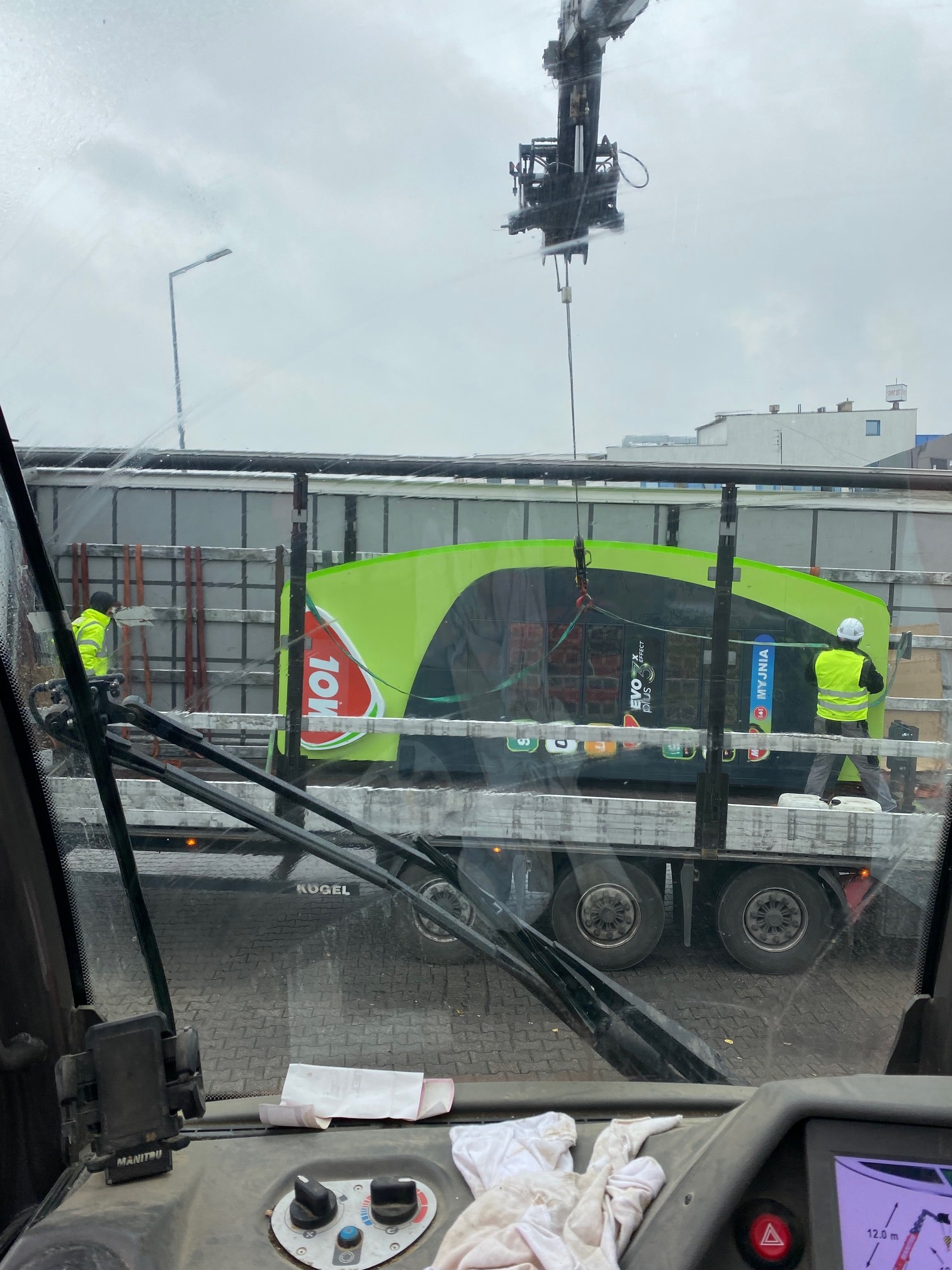 Widok z kabiny operatora na załadunek myjni samoobsługowej na platformę ciężarówki za pomocą dźwigu, pracownicy w kamizelkach odblaskowych zabezpieczają ładunek.