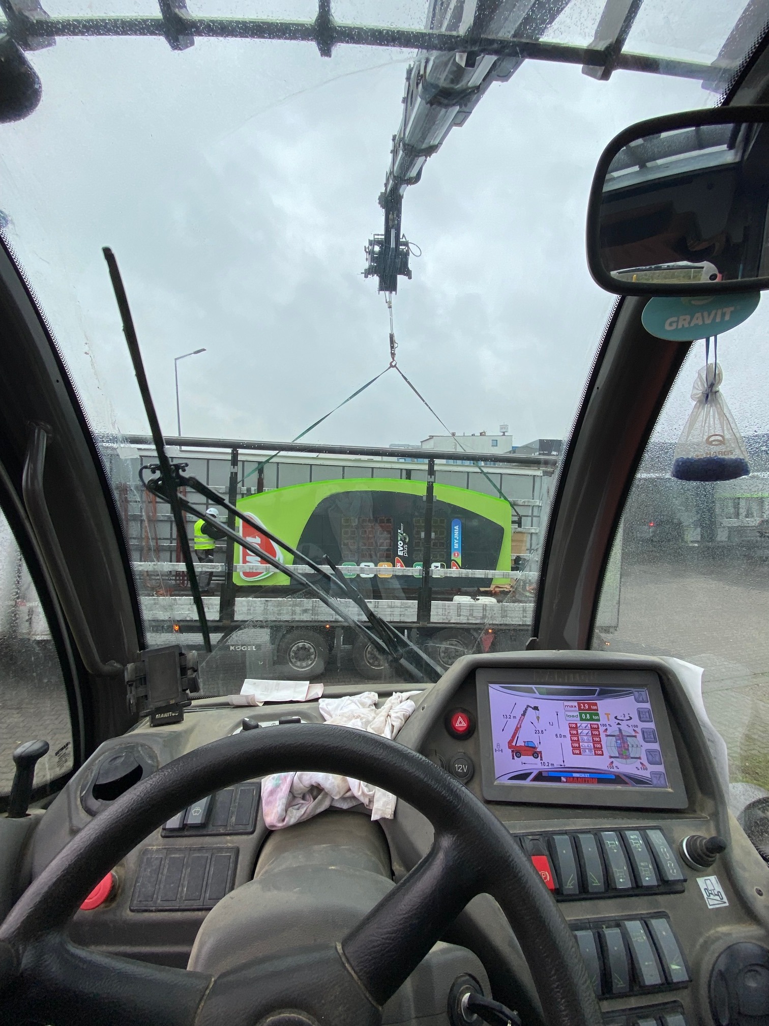 Widok z kabiny operatora żurawia samochodowego podczas załadunku zielonej myjni ciśnieniowej na naczepę ciężarówki. Na desce rozdzielczej widoczny ekran z parametrami pracy żurawia.