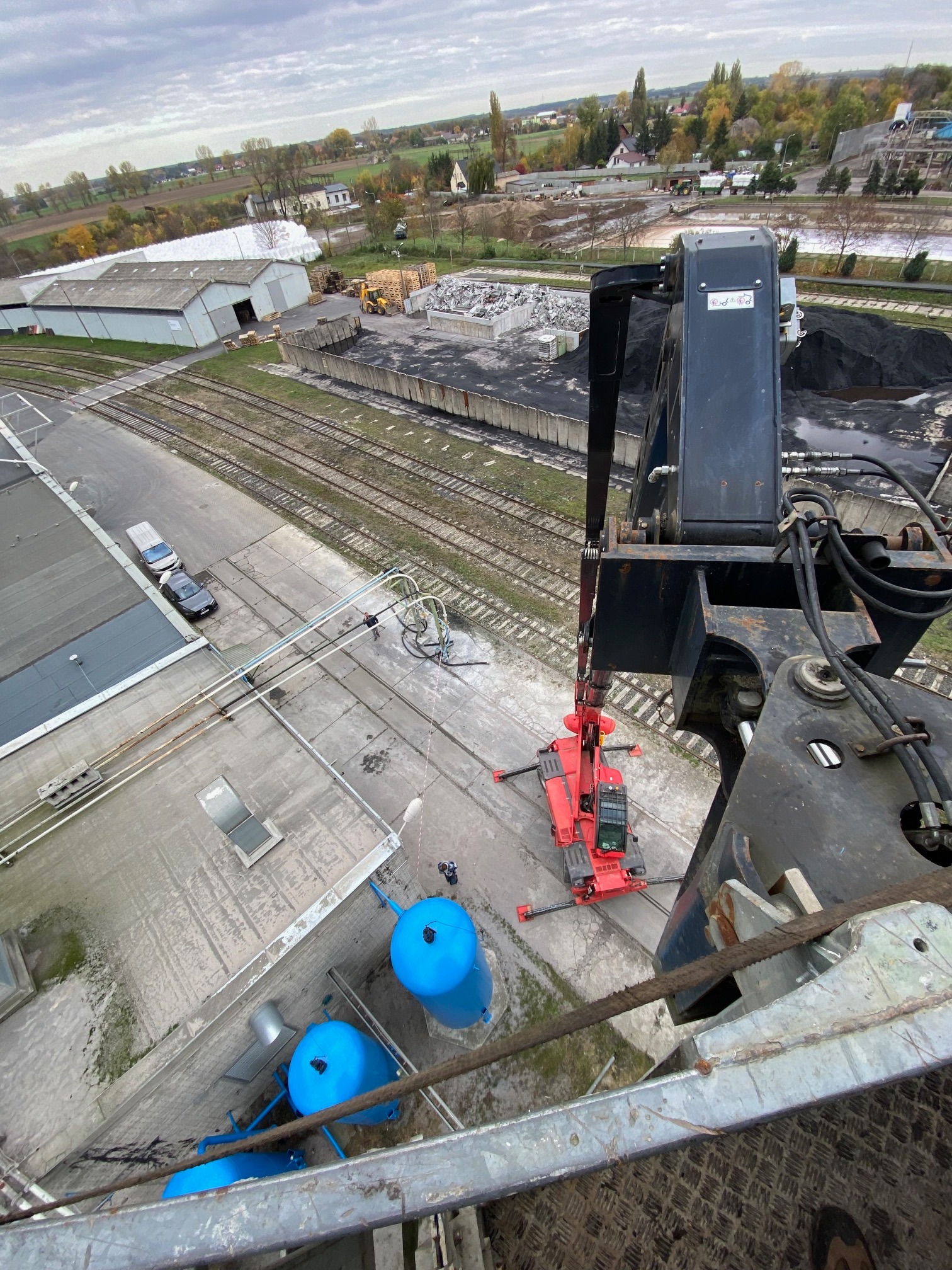Widok z góry na czerwoną miniładowarkę na betonowym placu, obok niebieskie zbiorniki, w tle budynki przemysłowe, torowisko i wieś.
