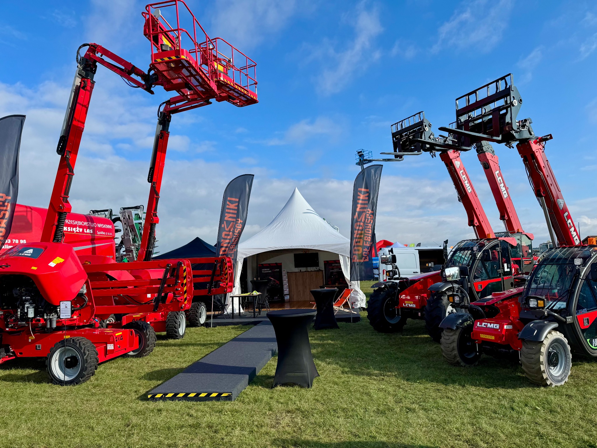 Czerwone ładowarki teleskopowe LOMG na trawie, z podnośnikiem koszowym i widłami, na tle białego namiotu i błękitnego nieba. Prezentacja maszyn na targach.
