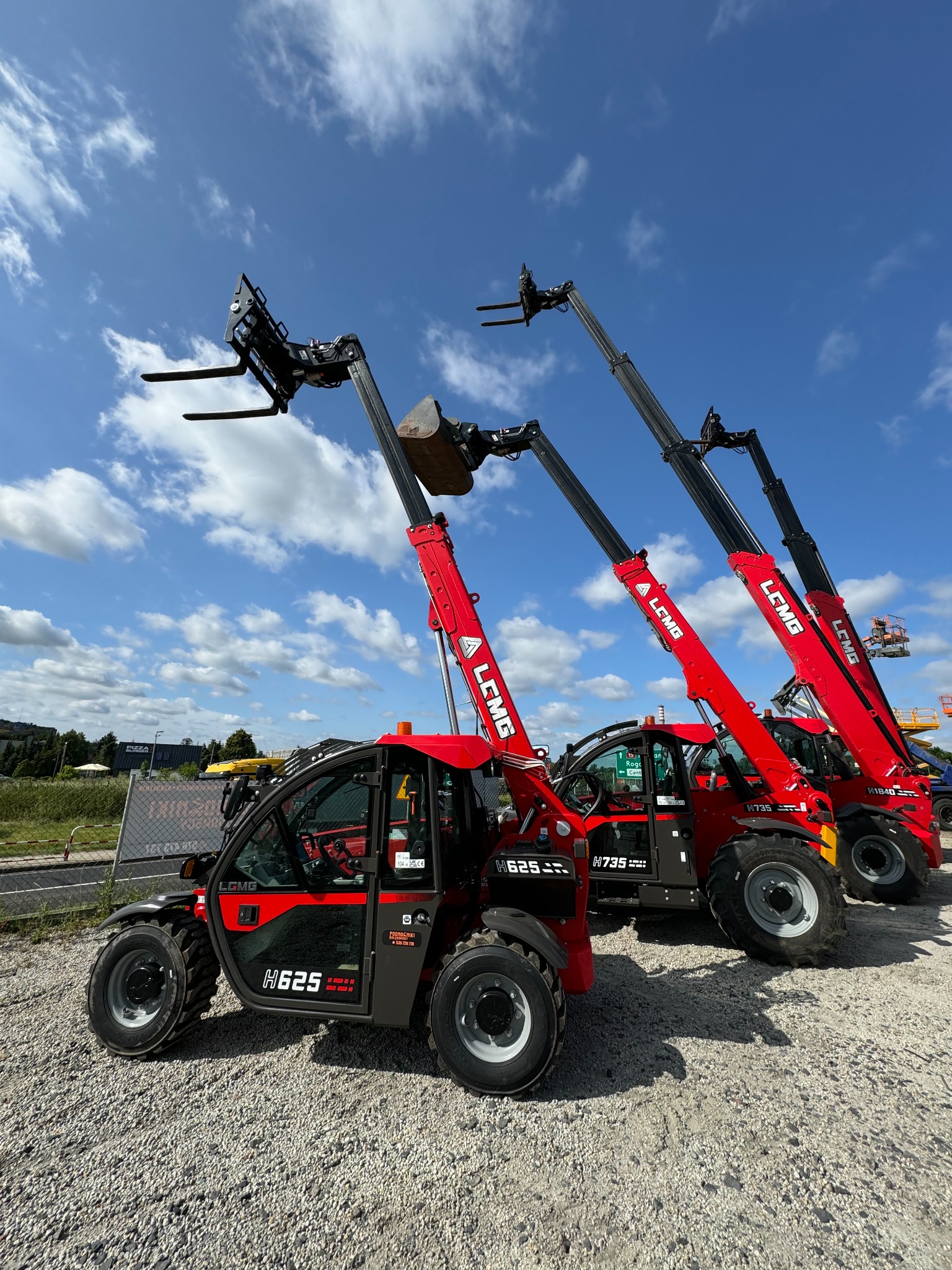 Rząd czerwonych ładowarek teleskopowych LGMG H625 i H735 na żwirowym placu, z podniesionymi ramionami i różnymi końcówkami, na tle błękitnego nieba z chmurami.