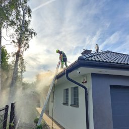 BLASK BRUK DOMINIK FIGZAŁ - Pracownik w Mstowie czyści dach myjką ciśnieniową, stojąc na drabinie opierającej się o rynnę, z widocznym efektem rozprysku wody i zanieczyszczeń.