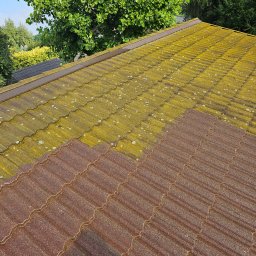 BLASK BRUK DOMINIK FIGZAŁ - Częściowo wyczyszczony dach z metalowej dachówki, widoczna różnica między obszarem pokrytym mchem i algami a czystą, czerwonobrązową powierzchnią. W tle zieleń drzew.