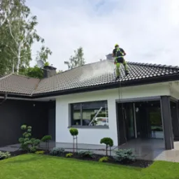 Pracownik w szelkach bezpieczeństwa czyści dach nowoczesnego domu z użyciem myjki ciśnieniowej, widoczny strumień wody i czyszczona powierzchnia dachu.