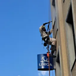 Pracownik w kombinezonie i kasku, zawieszony na linach, myje elewację budynku z logiem UNICA za pomocą myjki ciśnieniowej.