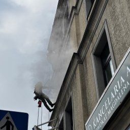 BLASK BRUK DOMINIK FIGZAŁ - Pracownik w uprzęży, myjący elewację budynku z wysokości, widoczny znak drogowy dla pieszych w lewym dolnym rogu i szyld 'Kancelaria Adwokat Magdalena A' po prawej stronie.