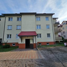 BLASK BRUK DOMINIK FIGZAŁ - Fasada bloku mieszkalnego z otwartymi drzwiami wejściowymi i kostką brukową przed wejściem, widoczne okna z roletami i balkony.