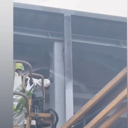 BLASK BRUK DOMINIK FIGZAŁ - Pracownik w kombinezonie maluje konstrukcję stalową natryskowo z podnośnika koszowego w Mstowie. Widoczne rozpylanie farby i elementy konstrukcji budynku.