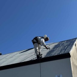 BLASK BRUK DOMINIK FIGZAŁ - Pracownik w kombinezonie maluje metalowy dach natryskowo, zabezpieczony linami. Widoczny adres budynku: ul. Nad Wartą 31, Łuszczyn. Błękitne niebo w tle.