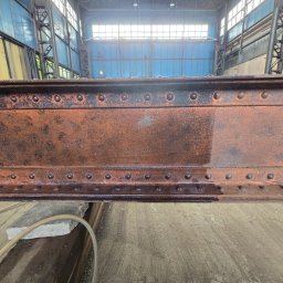 BLASK BRUK DOMINIK FIGZAŁ - Metalowa konstrukcja w hali przemysłowej, częściowo oczyszczona z rdzy. Widoczna różnica między stanem przed i po czyszczeniu, z detalami nitów i fakturą metalu.