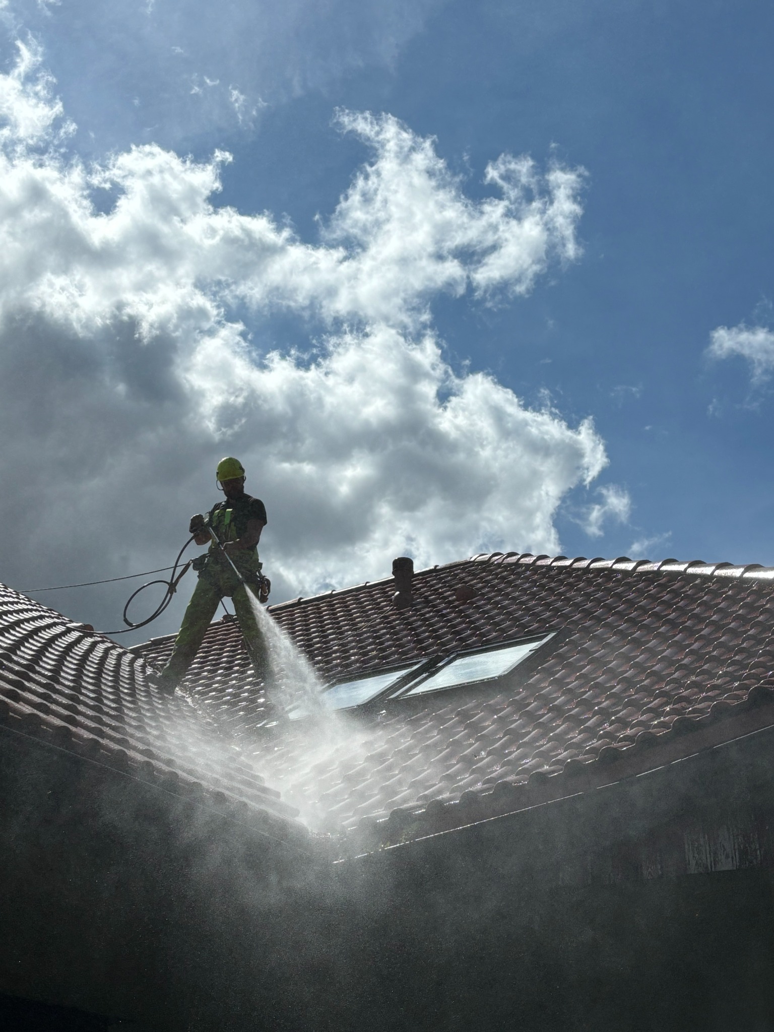 Pracownik w kasku i uprzęży, stojący na stromym, czerwonym dachu z okienkami dachowymi, czyści go za pomocą myjki ciśnieniowej na tle błękitnego nieba z chmurami.