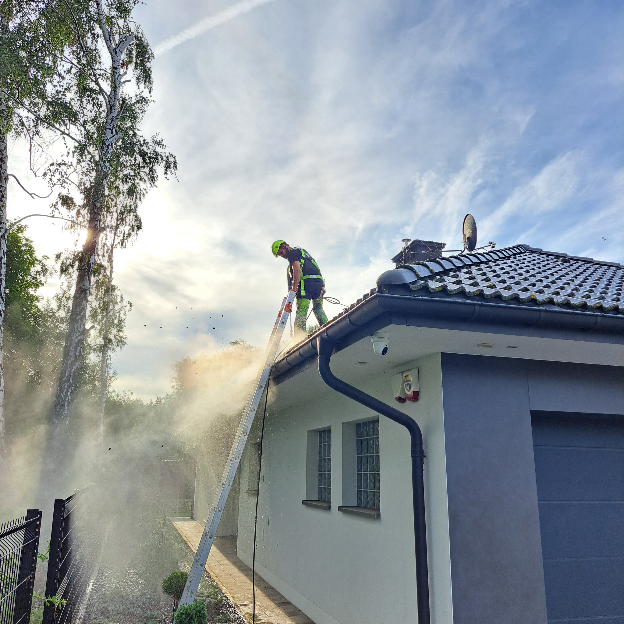 Pracownik w Mstowie czyści dach myjką ciśnieniową, stojąc na drabinie opierającej się o rynnę, z widocznym efektem rozprysku wody i zanieczyszczeń.