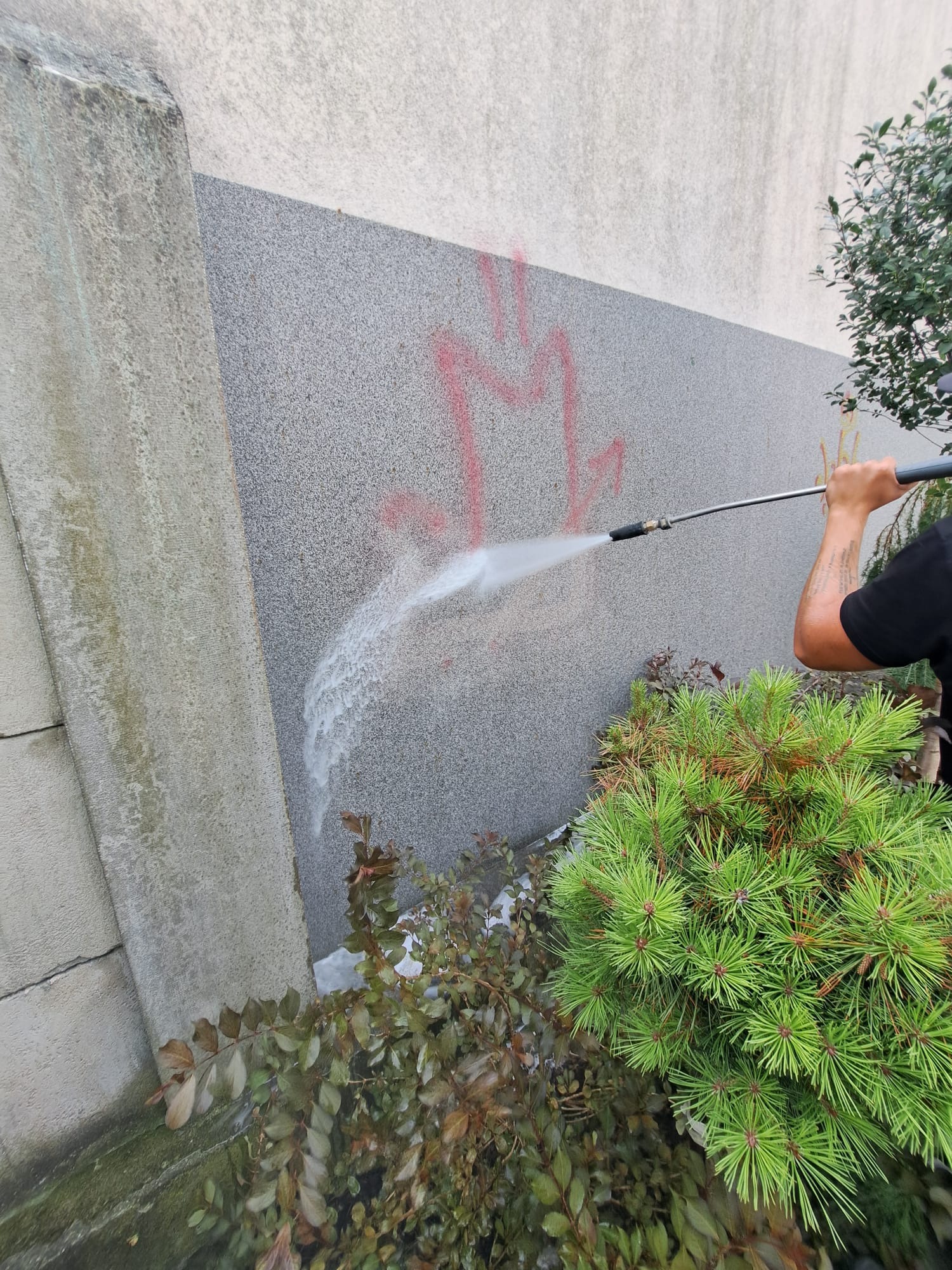 Czyszczenie szarej elewacji z czerwonego graffiti za pomocą myjki ciśnieniowej, widoczne rośliny w tle