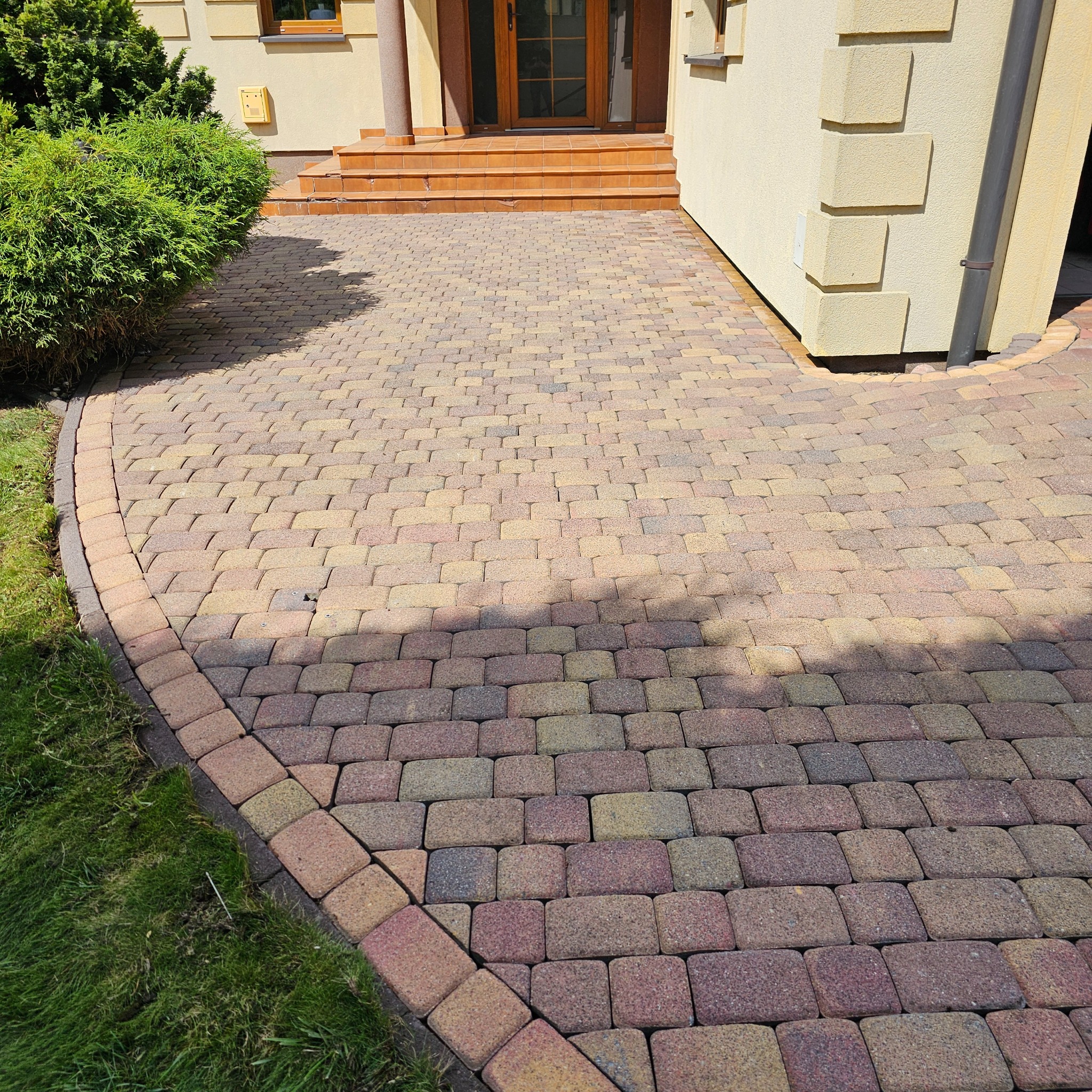 Ułożona kostka brukowa w odcieniach brązu i beżu tworzy ścieżkę prowadzącą do wejścia domu z zielonym trawnikiem po lewej stronie, widoczne schody wejściowe i fragment elewacji budynku.