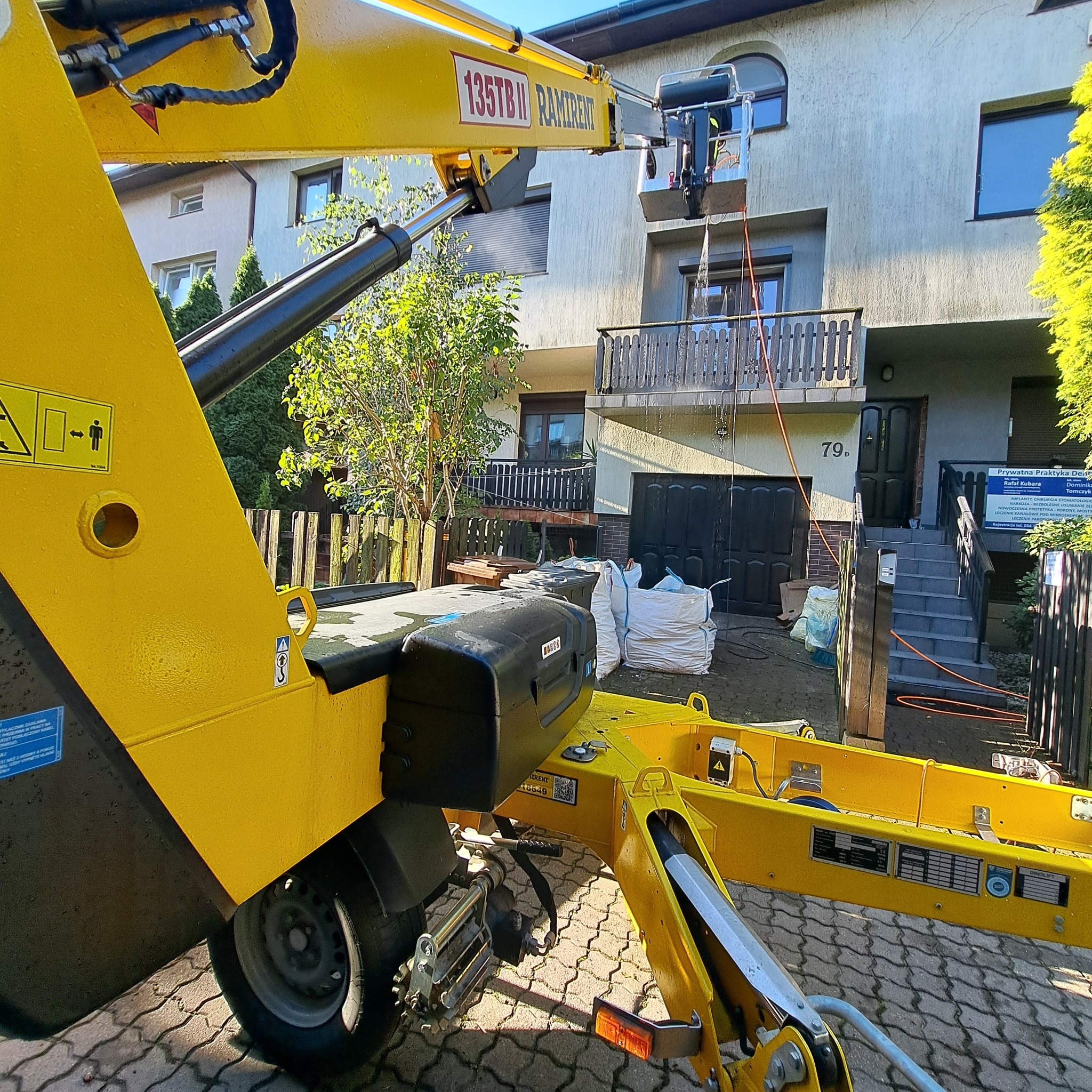 Żółty podnośnik koszowy marki Ramirent na chodniku z kostki brukowej, skierowany w stronę elewacji budynku z numerem 79d, w koszu pracownik podczas pracy, w tle widoczne worki z odpadami po remoncie.