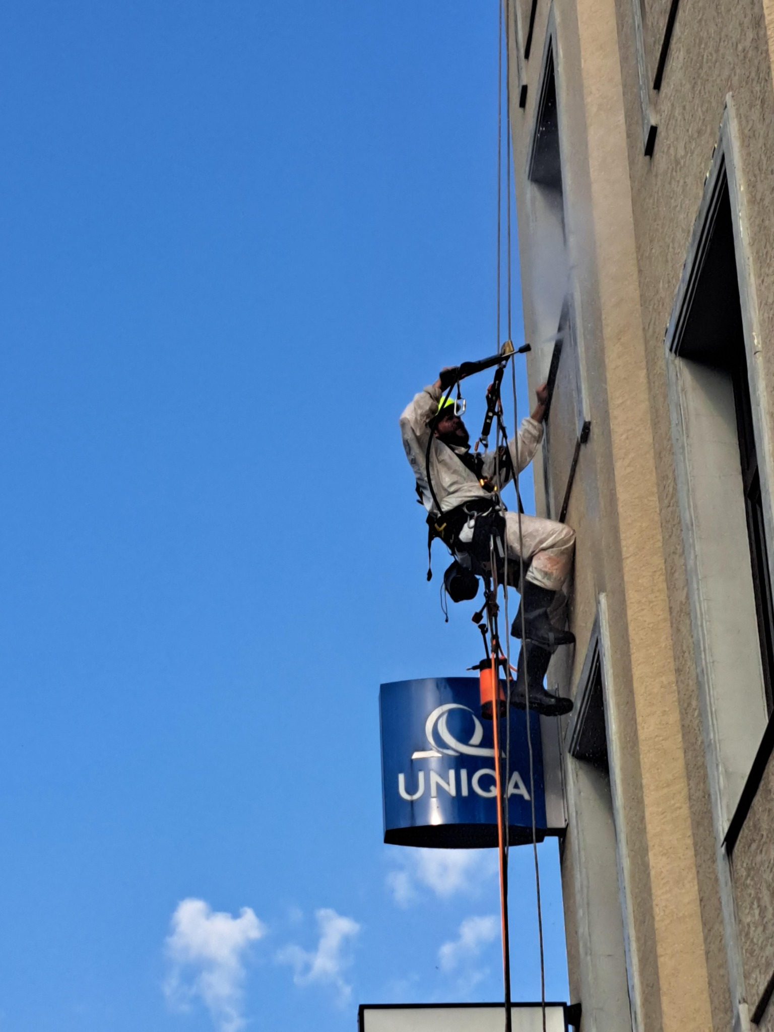 Pracownik w kombinezonie i kasku, zawieszony na linach, myje elewację budynku z logiem UNICA za pomocą myjki ciśnieniowej.
