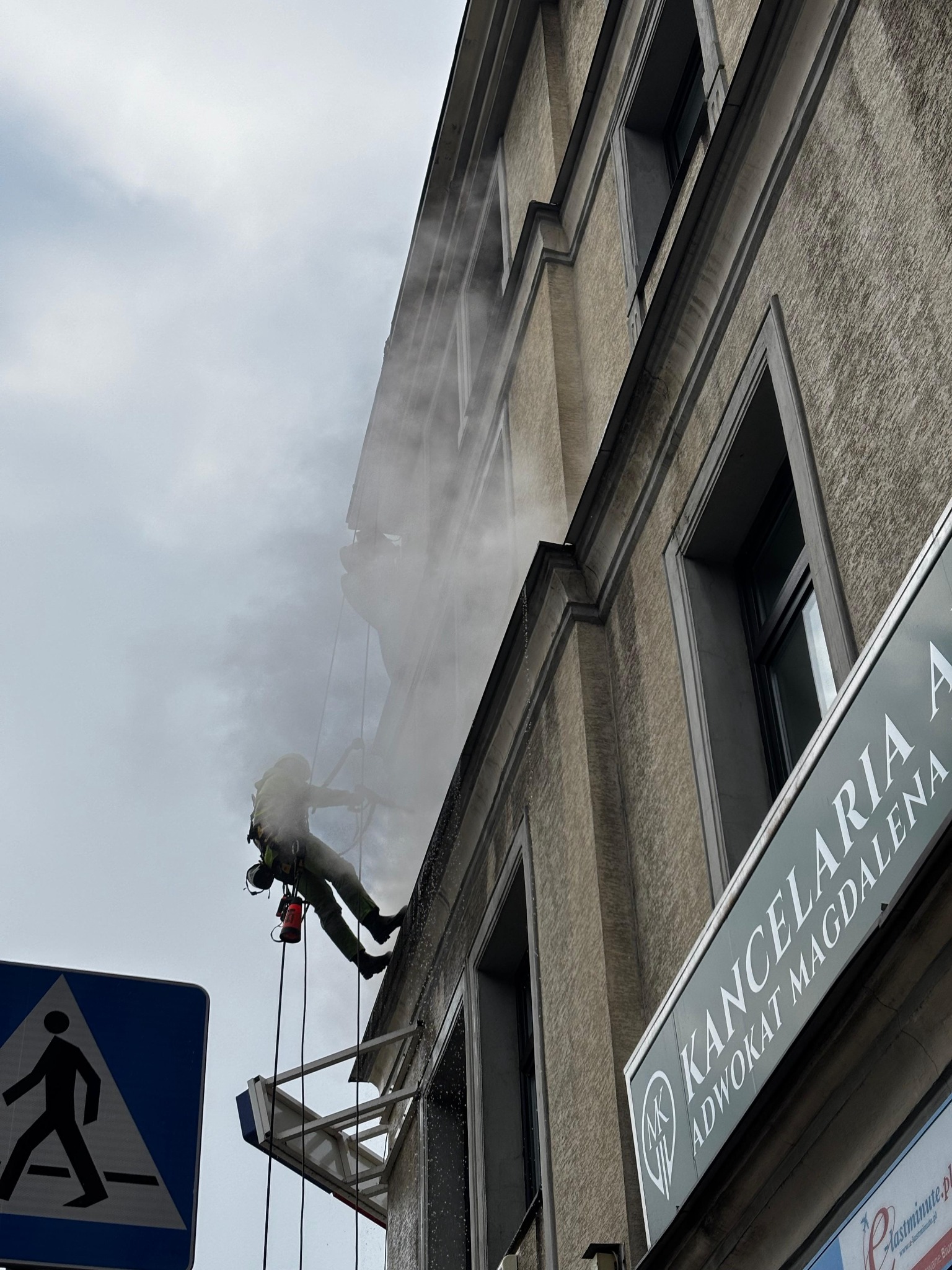 Pracownik w uprzęży, myjący elewację budynku z wysokości, widoczny znak drogowy dla pieszych w lewym dolnym rogu i szyld 'Kancelaria Adwokat Magdalena A' po prawej stronie.