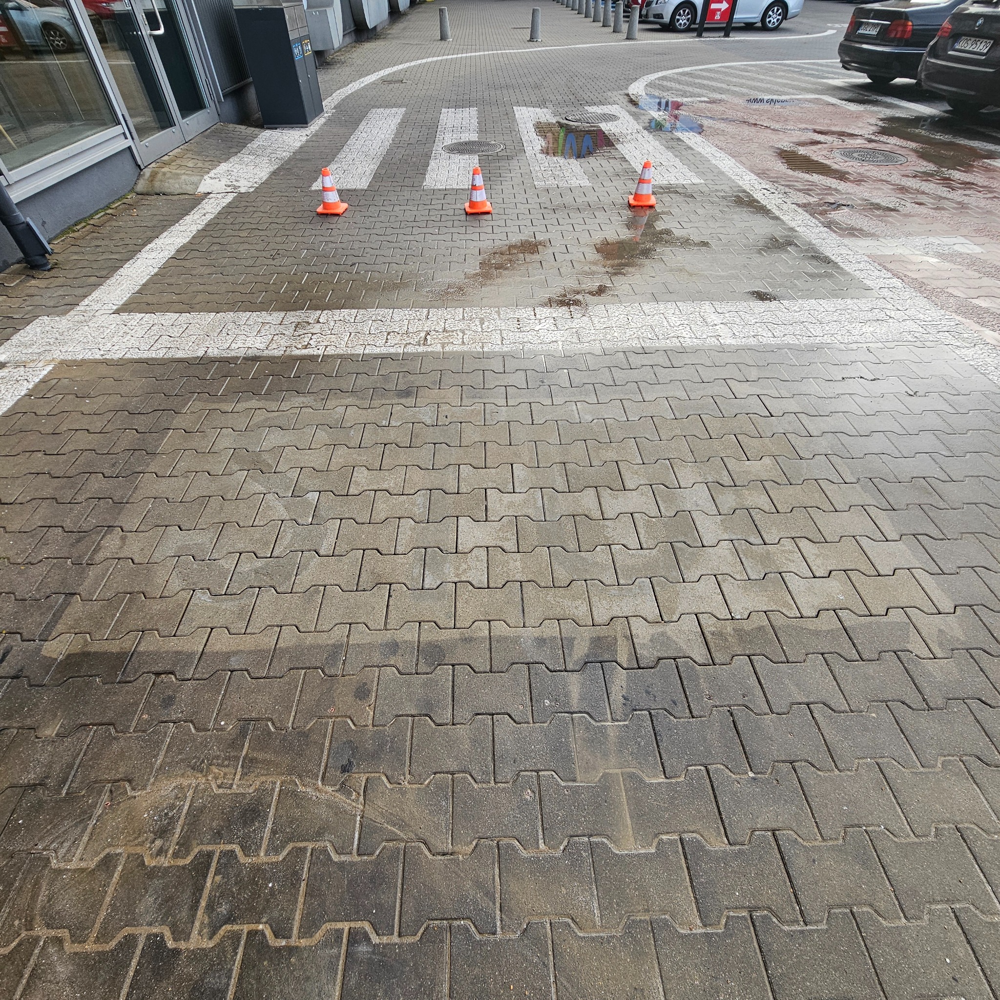Mokra kostka brukowa na przejściu dla pieszych z trzema pomarańczowymi pachołkami, widoczne ślady po czyszczeniu, w tle fragment parkingu i budynek.
