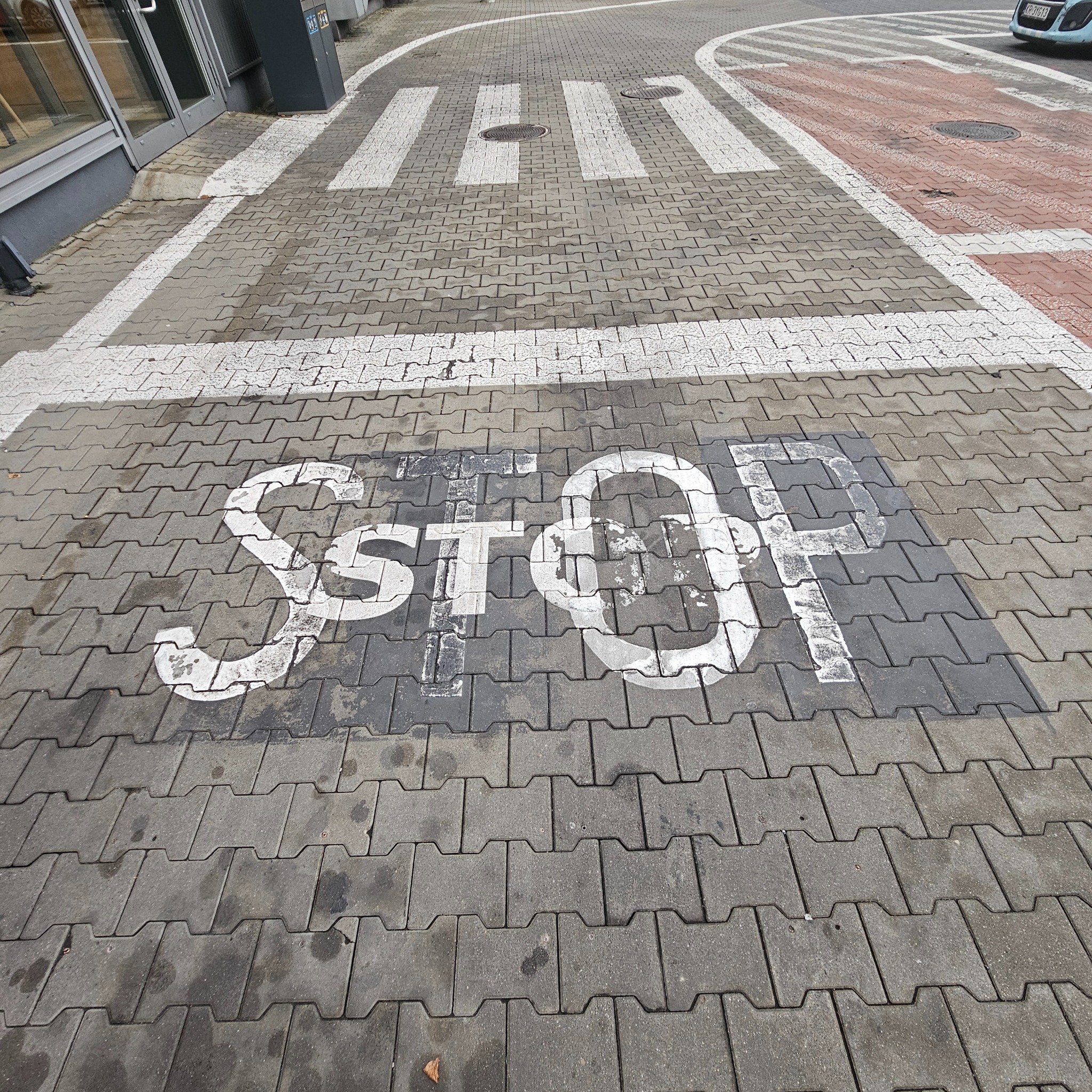 Szara kostka brukowa z namalowanym białym napisem STOP i przejściem dla pieszych w tle, widoczne ślady użytkowania.