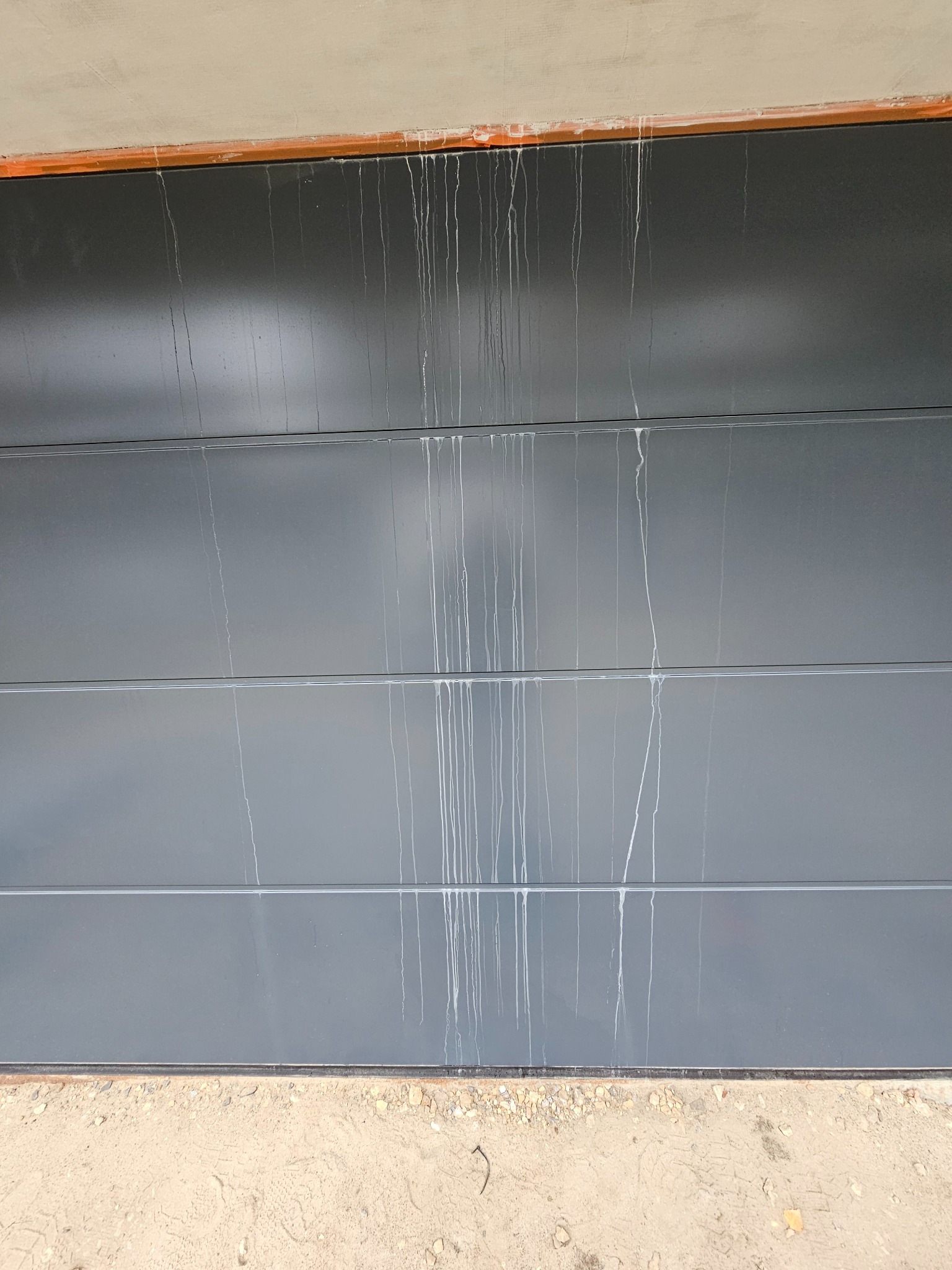 Szara, segmentowa brama garażowa z zaciekami po nieudanym czyszczeniu, widoczne ślady spływającego płynu, piasek u podstawy bramy.