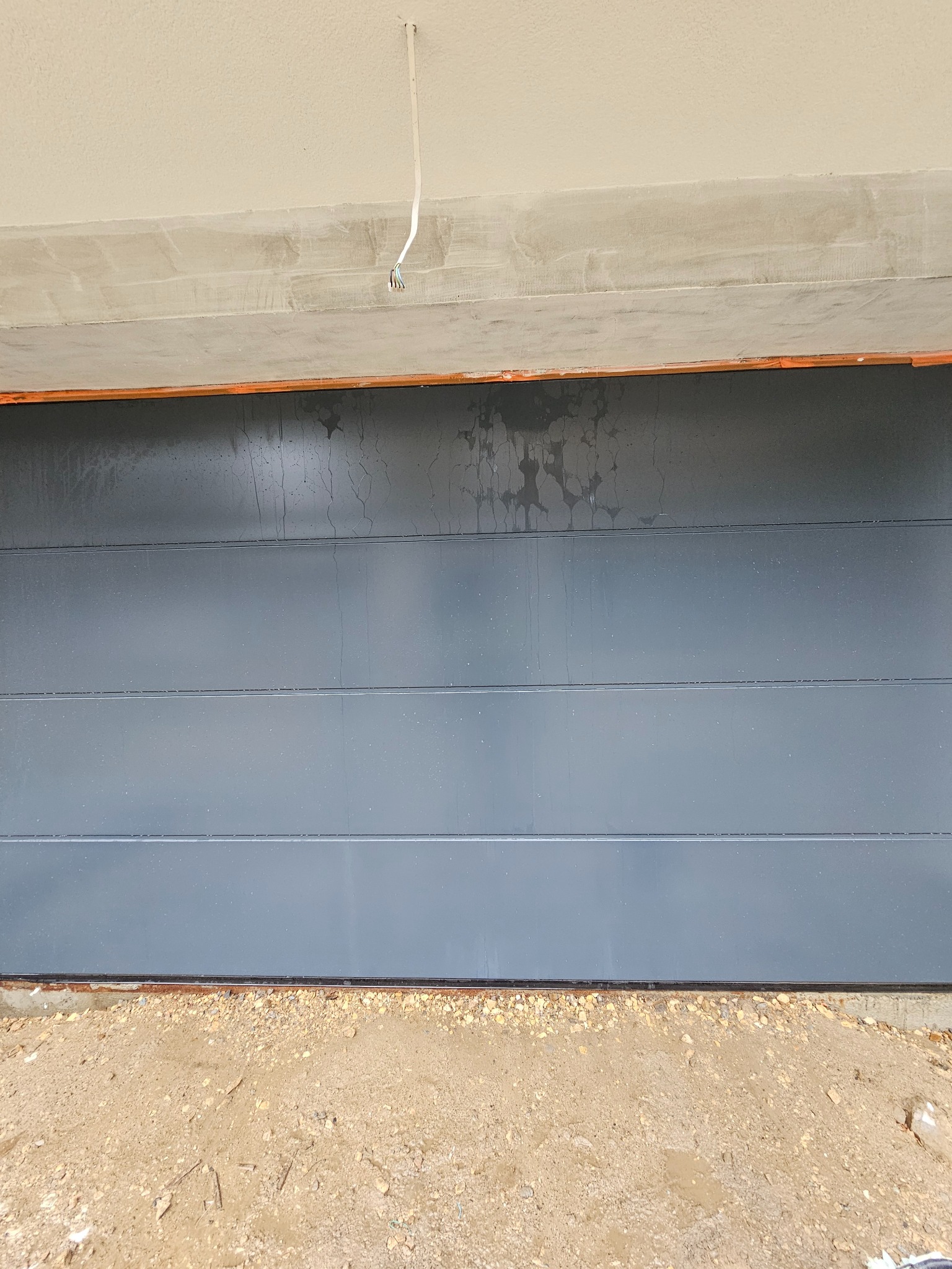 Szara, segmentowa brama garażowa w trakcie montażu, widoczne ślady wody i niedokończone wykończenie sufitu oraz podłoże z piasku i kamieni.