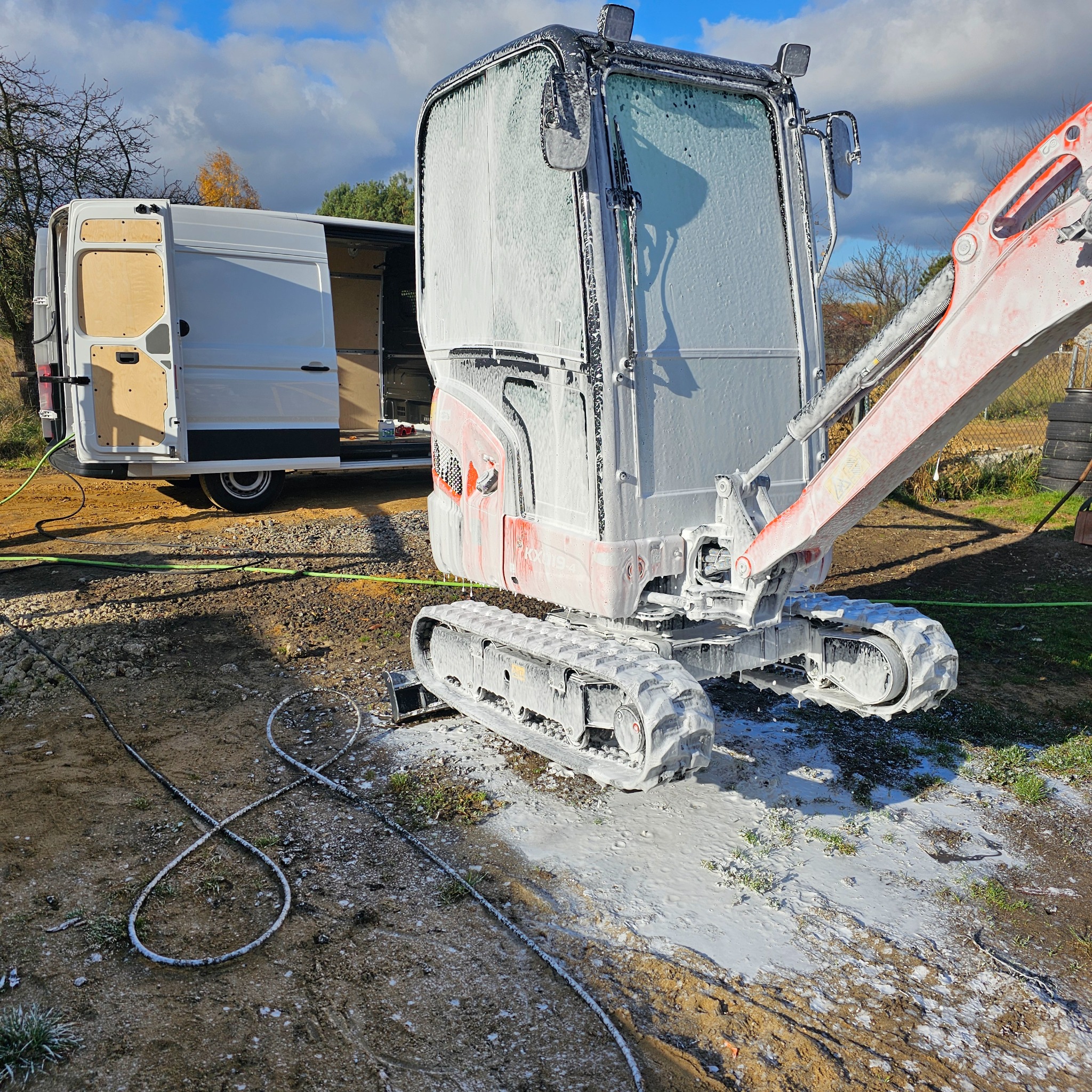 Mini koparka pokryta pianą czyszczącą, zaparkowana obok białego vana z otwartymi drzwiami, na ziemi widoczne kable i resztki piany.