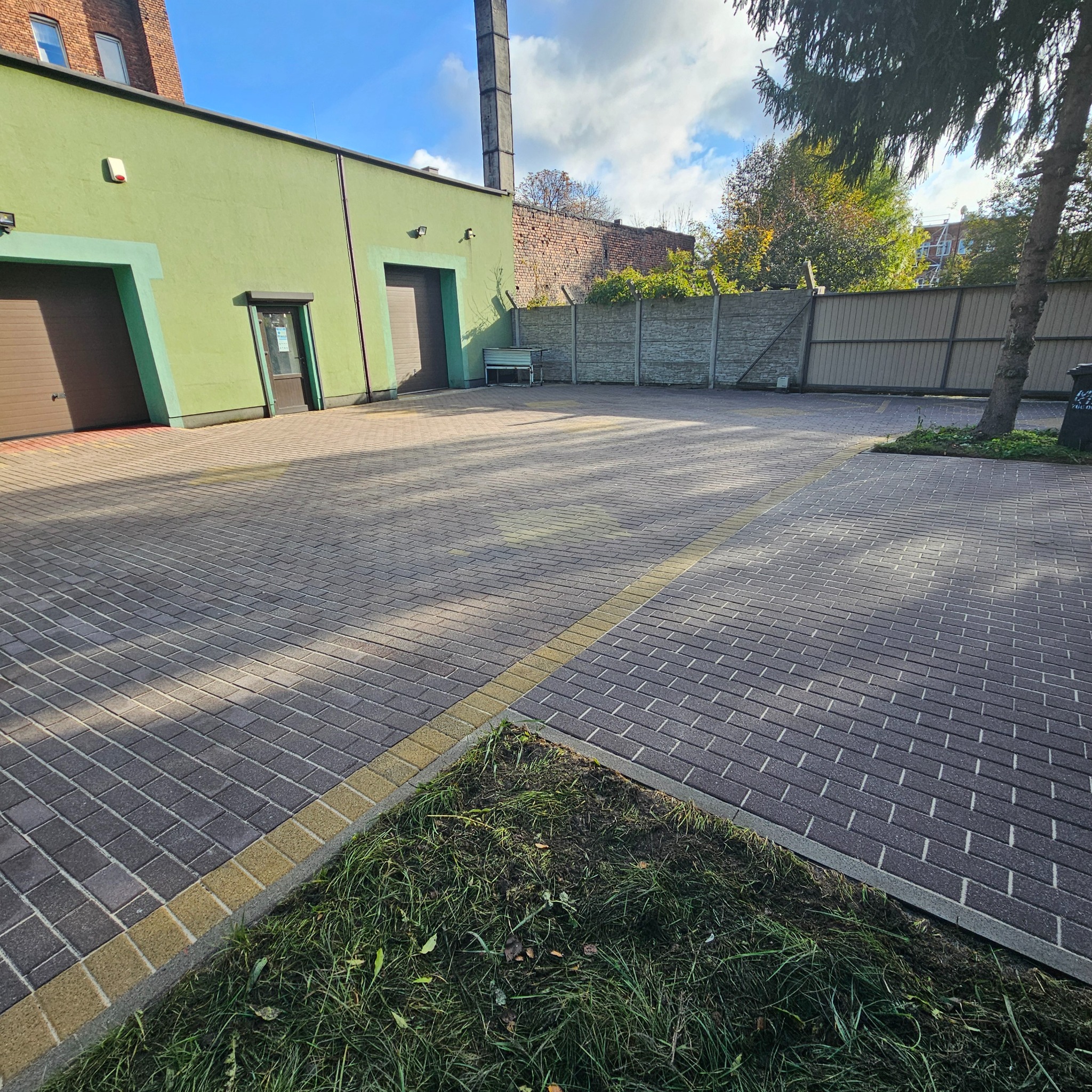 Nowo ułożona kostka brukowa w odcieniach szarości z żółtym obrzeżem na podjeździe przed budynkiem z zieloną elewacją i brązowymi bramami garażowymi.