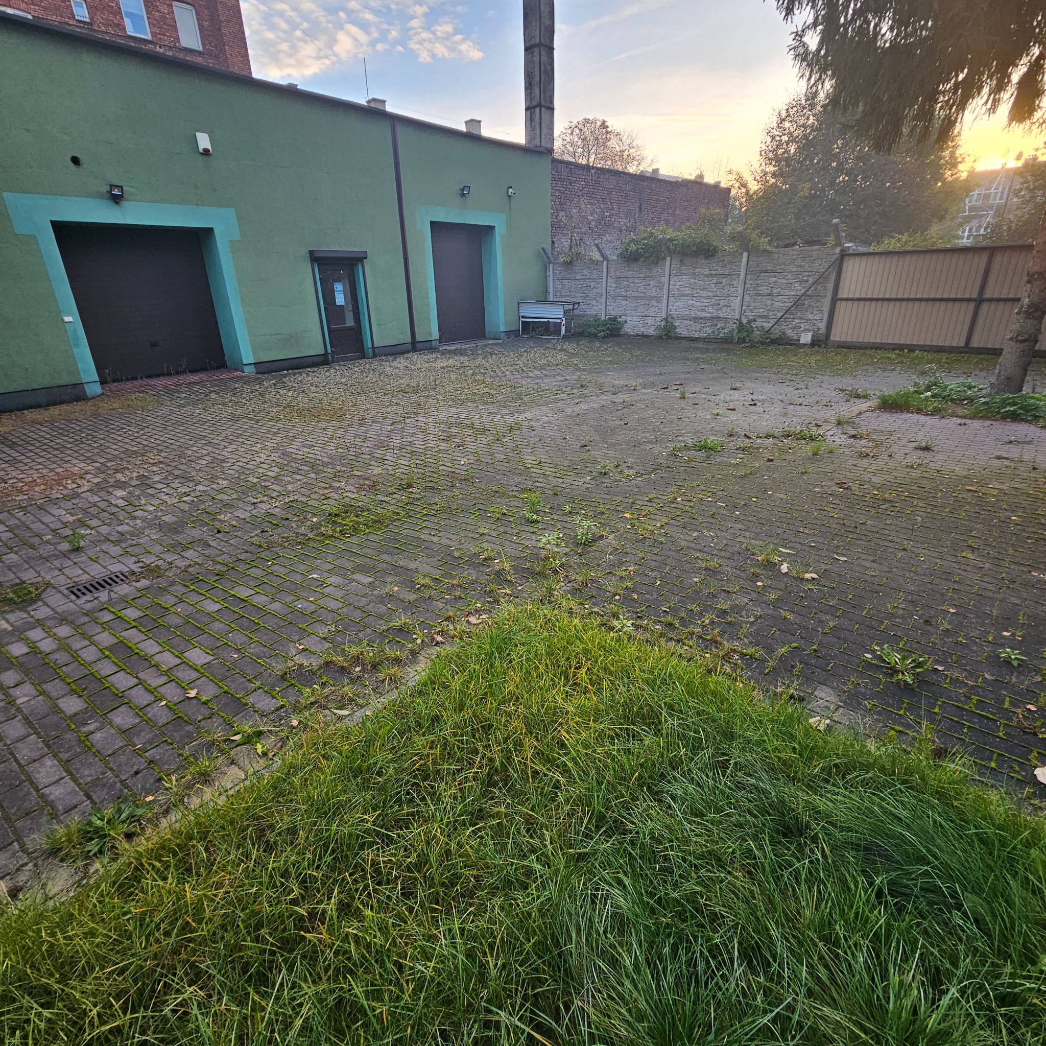 Zaniedbany plac z kostki brukowej porośniętej mchem i trawą, z zielonym budynkiem z dwoma bramami garażowymi w tle, widoczne pozostałości liści i zanieczyszczeń.