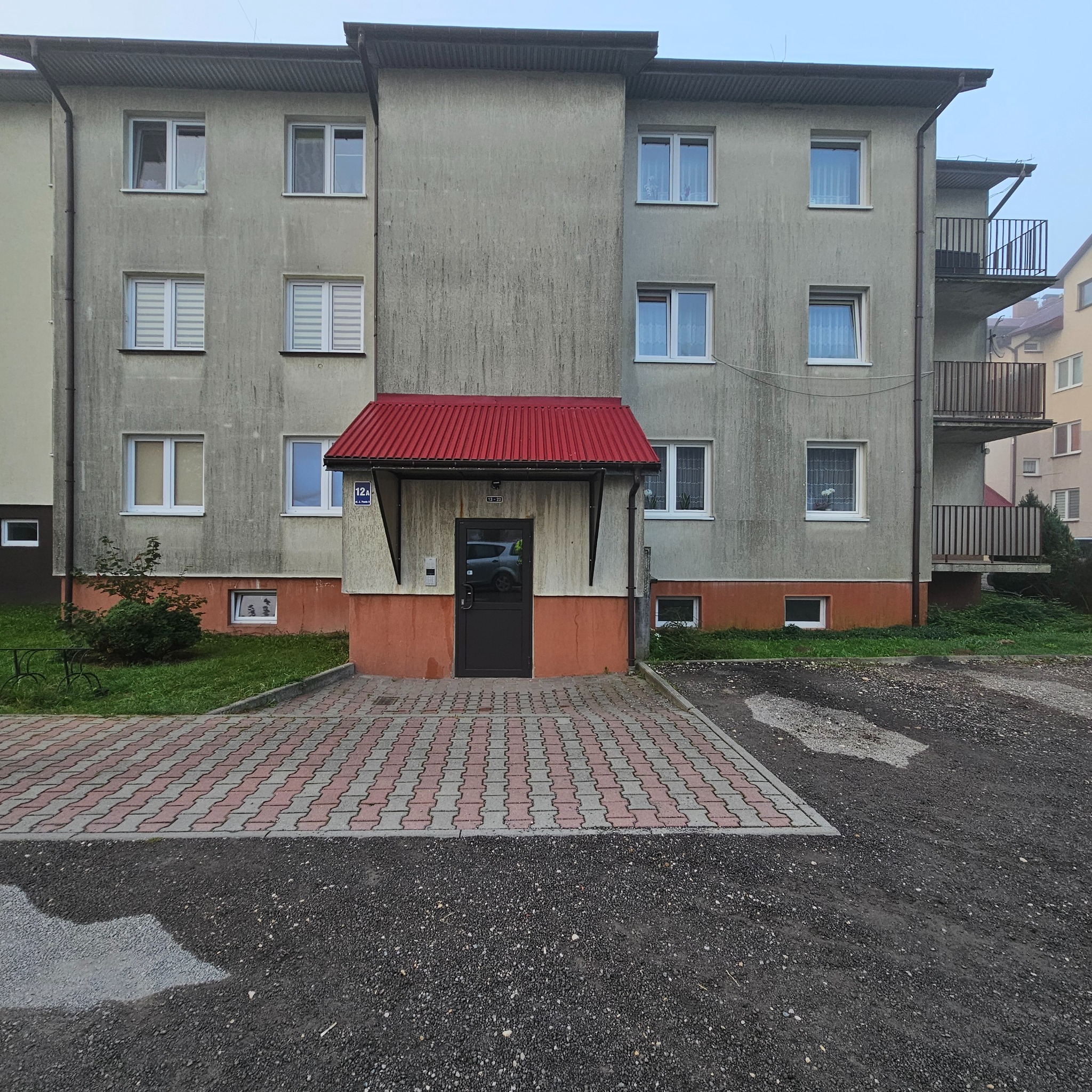 Szary blok mieszkalny z czerwonym zadaszeniem nad wejściem, widoczne ślady zabrudzeń na elewacji.