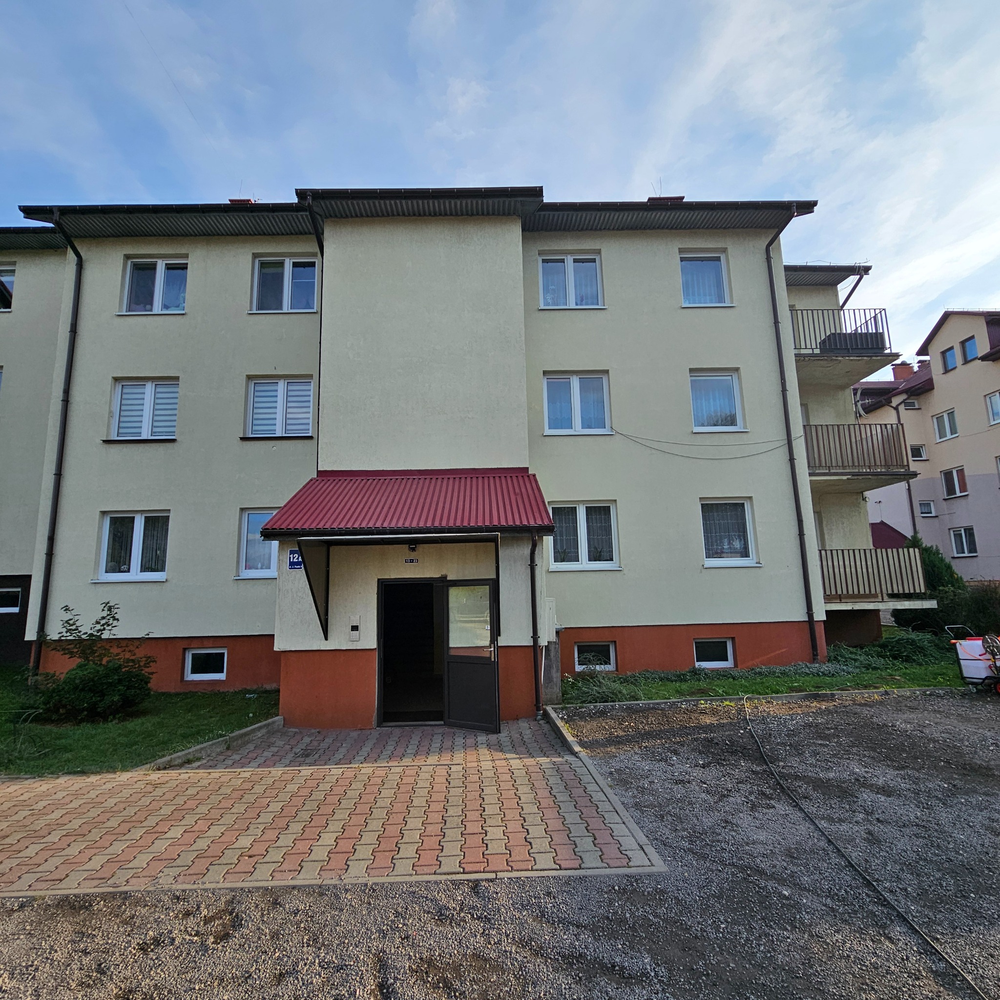 Fasada bloku mieszkalnego z otwartymi drzwiami wejściowymi i kostką brukową przed wejściem, widoczne okna z roletami i balkony.