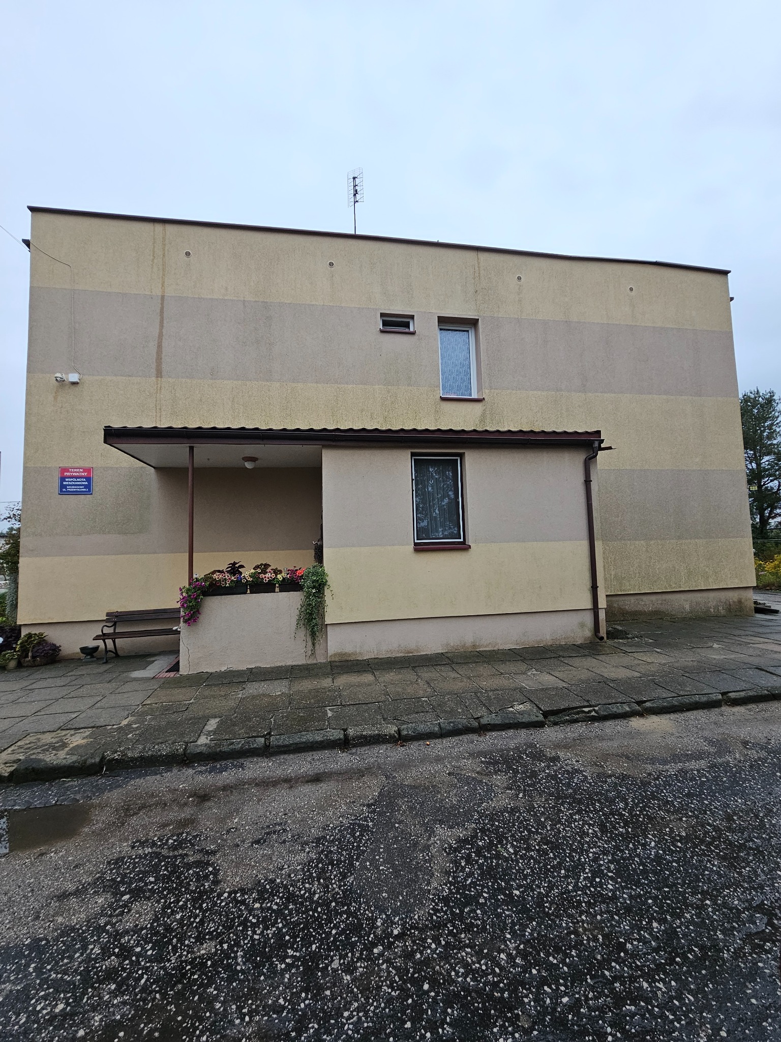 Dwukondygnacyjny budynek z elewacją w odcieniach beżu i szarości, widoczne okna, zadaszone wejście z kwiatami w donicach, ławka i tabliczka informacyjna na ścianie, mokry chodnik i jezdnia przed...