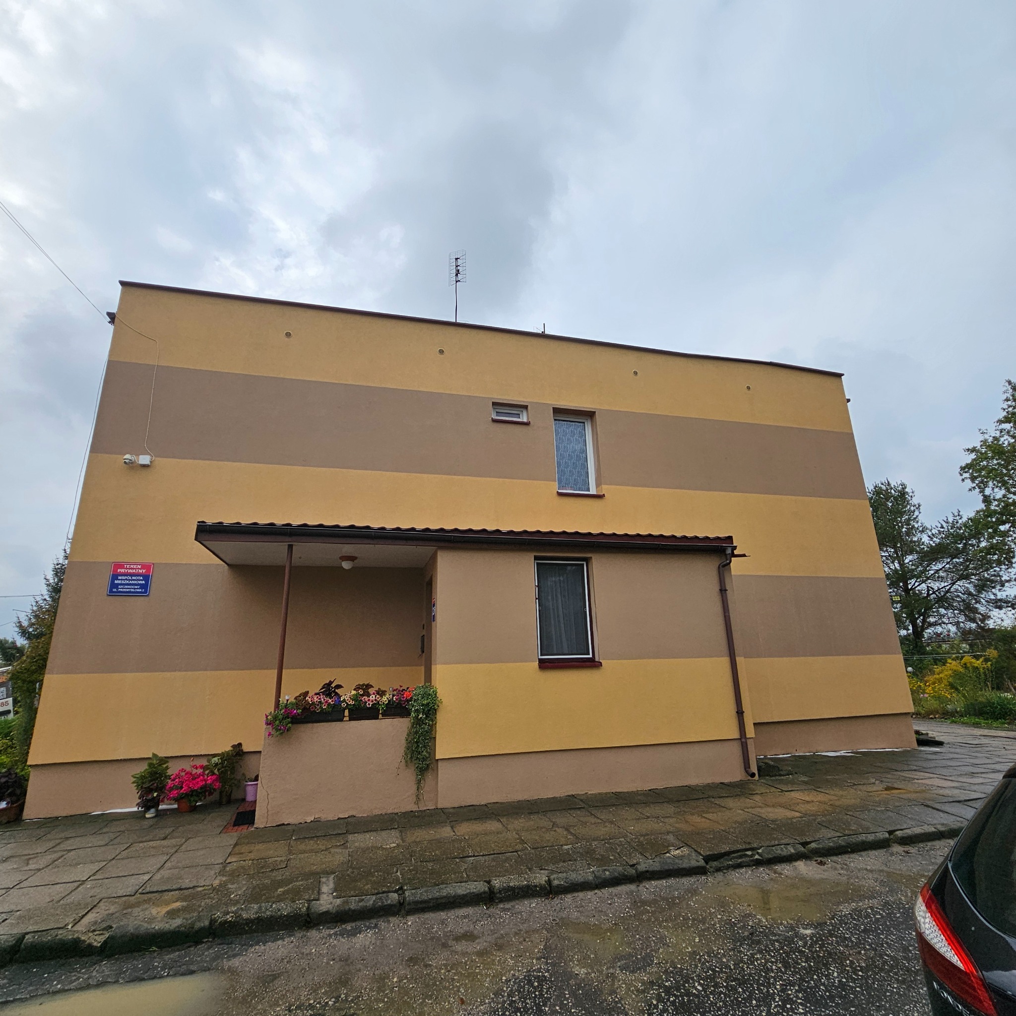 Dwukondygnacyjny budynek z elewacją w poziome pasy brązu i beżu, widok z dołu w pochmurny dzień, z widocznymi oknami i kwiatami w donicach przy wejściu.