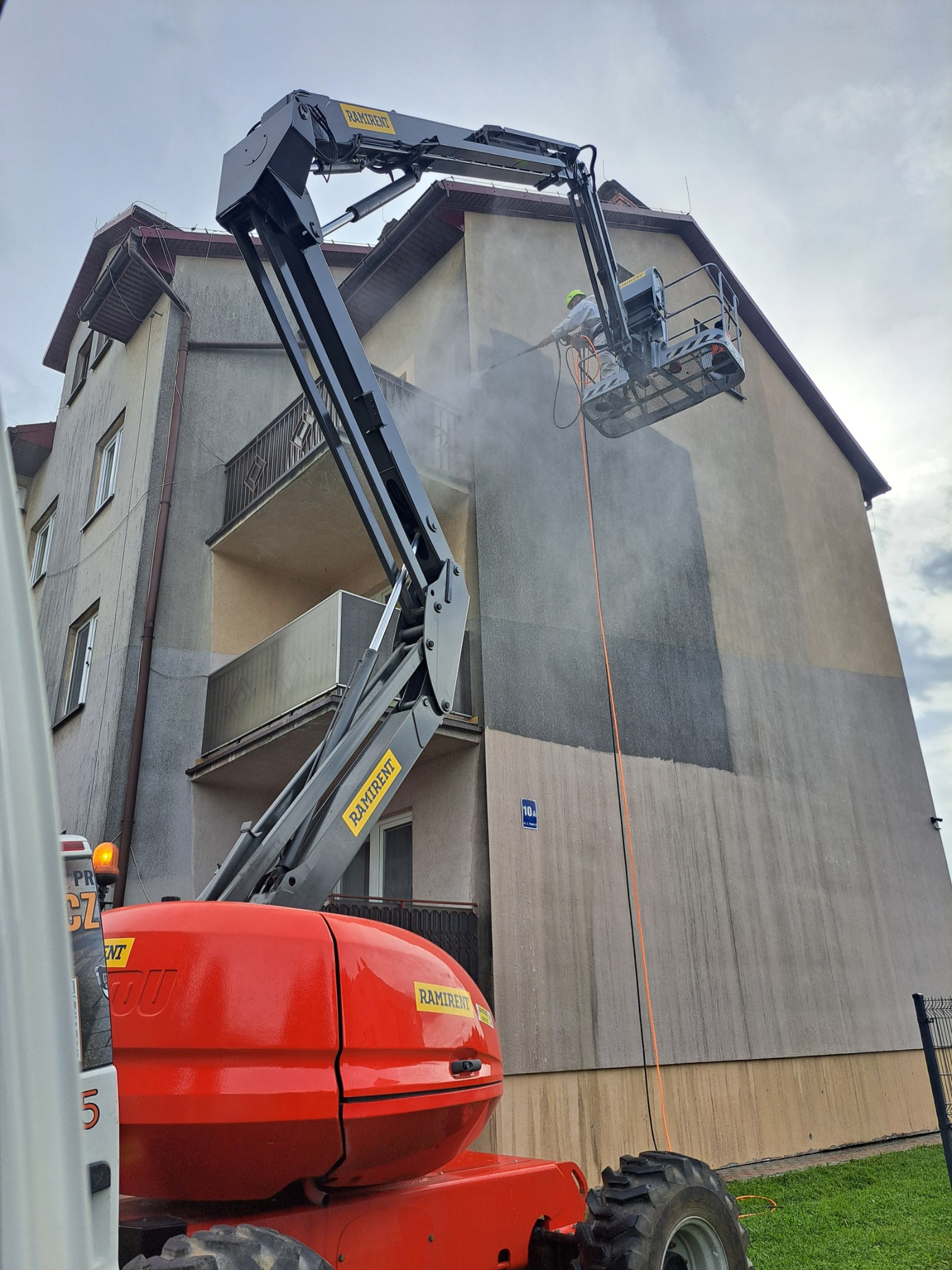 Pracownik w kombinezonie ochronnym na podnośniku koszowym czyści elewację budynku wielorodzinnego za pomocą myjki ciśnieniowej, widoczne różnice w kolorze elewacji przed i po czyszczeniu.